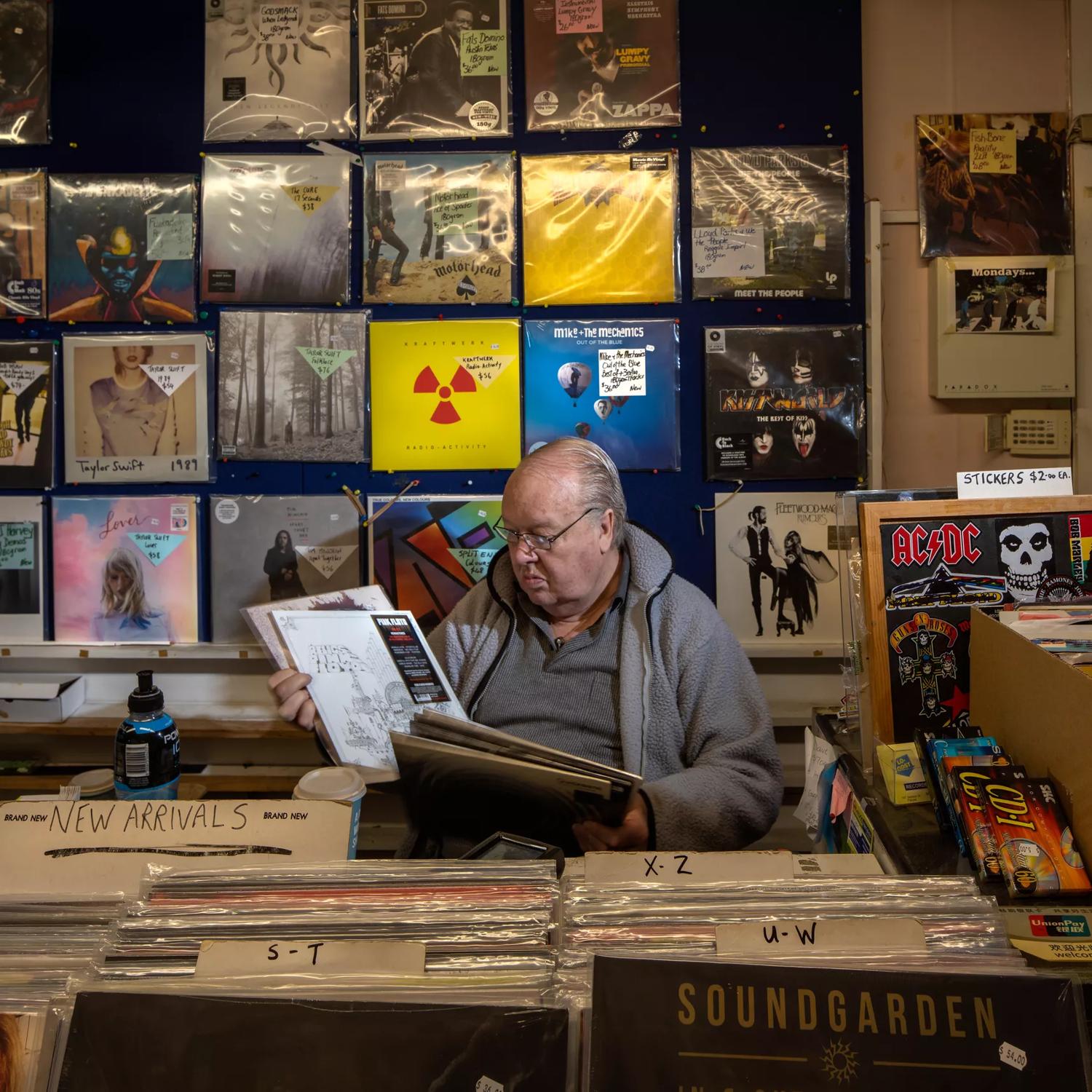 A man sits behind the counter looking through a stack of vinyls at Lo-Cost / Moonhop, a music store in Petone, Lower Hutt, New Zealand. 