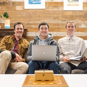 Three people sit on a couch holding laptops, smiling at the camera.