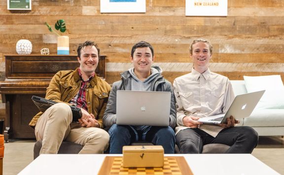 Three people sit on a couch holding laptops, smiling at the camera.