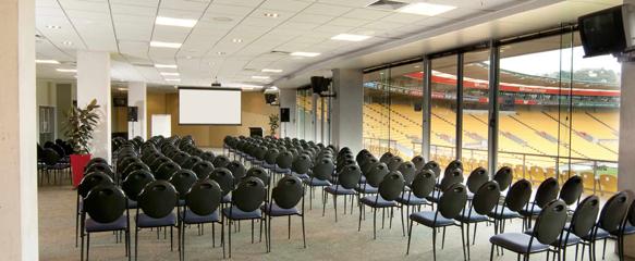 Inside the Level 2 East and West Lounges at Hnry Stadium Function Centre, with roughly 100 chairs facing toward a projector. The rugby field is on the right, outside floor-to-ceiling windows.