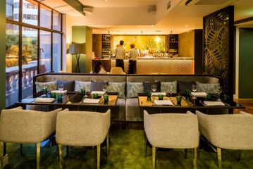 Inside the DoubleTree by Hilton Wellington restaurant and bar. Cream chairs sit at square café tables with grey bench seats. Serving staff stand at the golden bar in the distance.