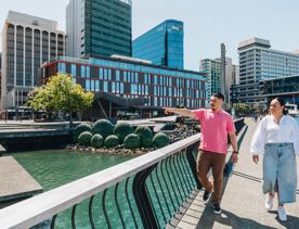 Two people are standing on Kumutoto Wharf near Lambton Quay on a sunny day. The water behind them is green and the sky is blue.