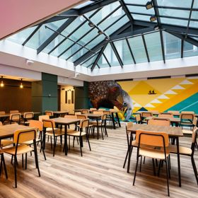 A shared dining space inside the Haka House hostel on Wakefield Street. A large mural of a Kākā is on the wall, and the roof is glass.