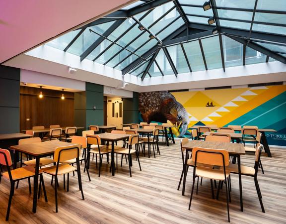 A shared dining space inside the Haka House hostel on Wakefield Street. A large mural of a Kākā is on the wall, and the roof is glass.