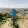 Dirt track with a blue sign post that has text 'Rough Justice' on it. Rolling hills and a city can be seen in the background.