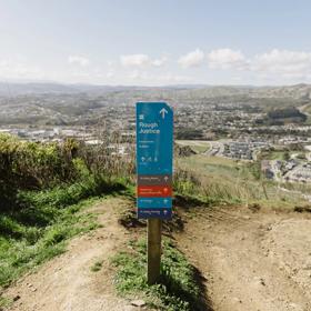 Dirt track with a blue sign post that has text 'Rough Justice' on it. Rolling hills and a city can be seen in the background.