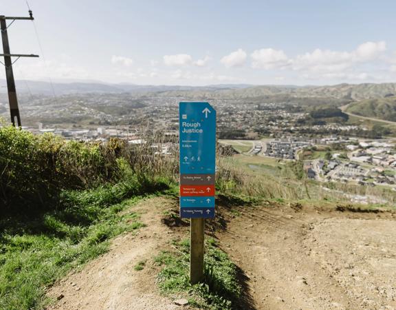 Dirt track with a blue sign post that has text 'Rough Justice' on it. Rolling hills and a city can be seen in the background.