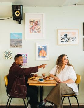 Two people sit at a table inside Swimsuit on Dixon Street, talking and drinking coffee. The wall behind them is duck egg green, and the wall is covered in art.