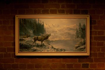 A framed painting of a landscape with pine trees, a lake, two moose and mountains affixed to a brick wall at Dee's Place, a cocktail bar in Wellington.