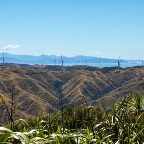 A scenic view from the Mākara Peak Mountain Bike Park toward the South Island with twelve wind turbines standing along a mountain range and blue skies above.