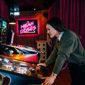 The interior of Mean Doses Taproom, a Brewery and Fillery located at 66 Tory Street, Te Aro, Wellington, with two vintage pinball machines and a pink neon sign in the background. A person wearing a green jumper is playing the pinball machine in the foregr