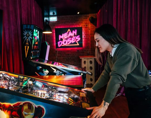 The interior of Mean Doses Taproom, a Brewery and Fillery located at 66 Tory Street, Te Aro, Wellington, with two vintage pinball machines and a pink neon sign in the background. A person wearing a green jumper is playing the pinball machine in the foregr