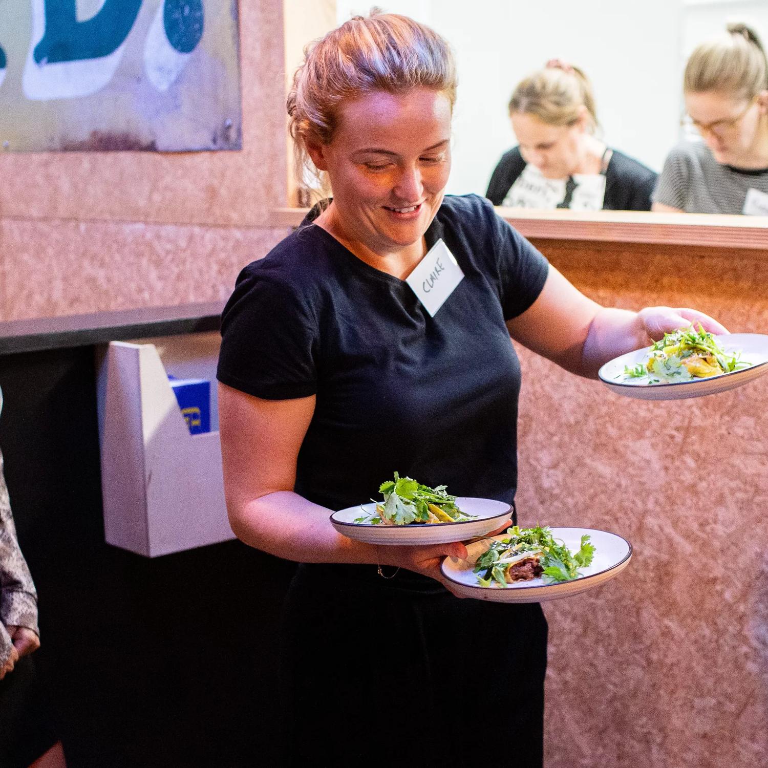 A person picks up three plates from the kitchen to deliver to tables at Everybody Eats in Wellington.