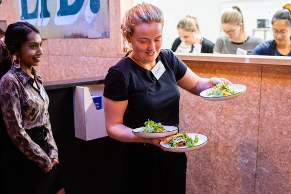 A person picks up three plates from the kitchen to deliver to tables at Everybody Eats in Wellington.