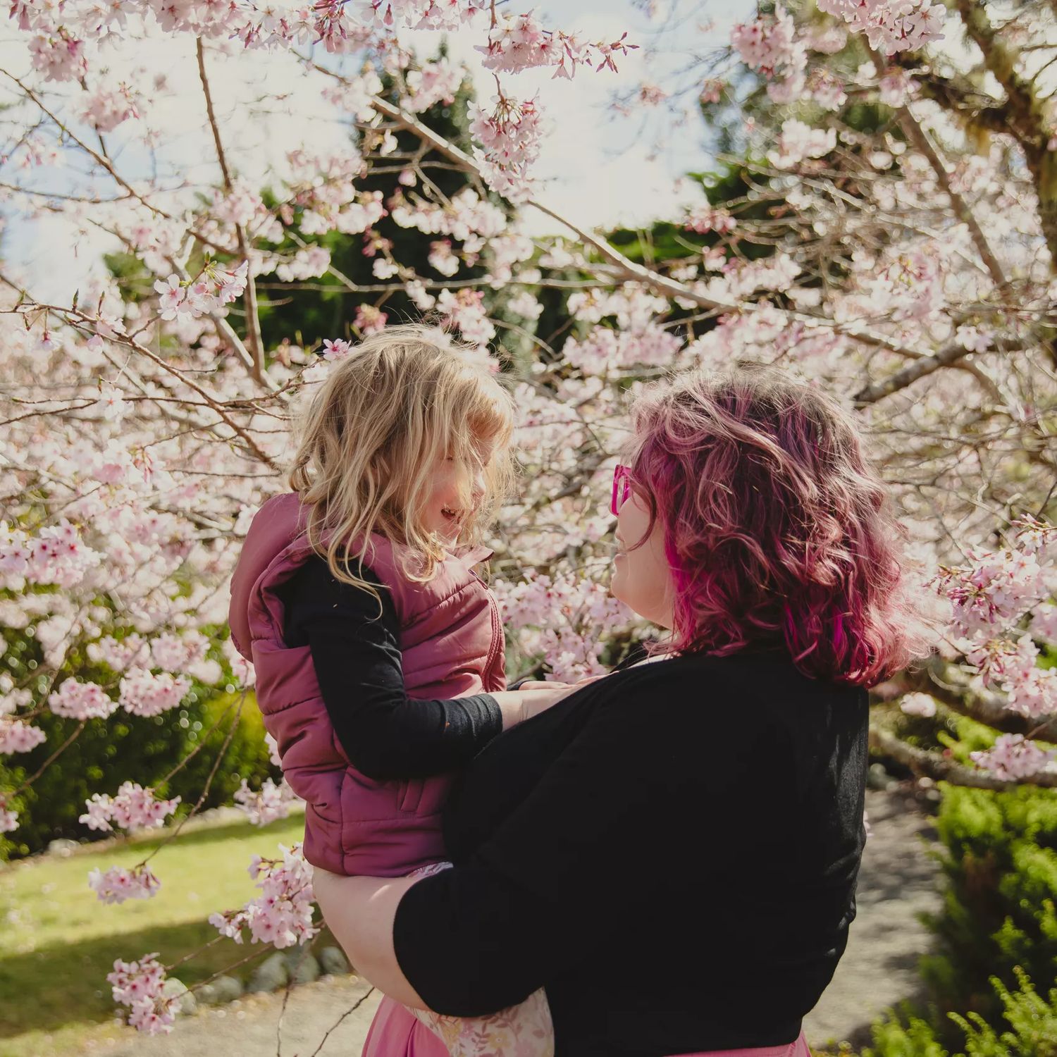 A parent holds their child in their arms with cherry blossom trees in bloom behind them.
