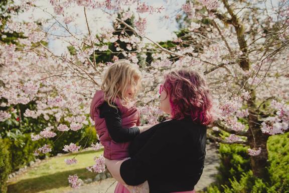 A parent holds their child in their arms with cherry blossom trees in bloom behind them.