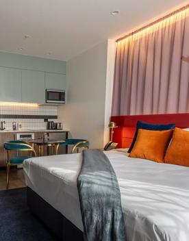 Premium King guest room at the Tryp by Wyndham. A bed with crisp white sheets and colourful cushions sits against the wall, with a kitchenette and café table and chairs in the background.