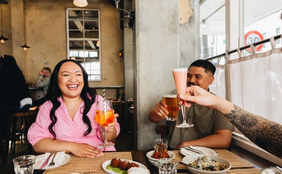 Three people cheers their cocktails over their food and laugh inside Ombra on Cuba Street.