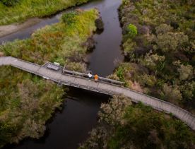 A drone shot of two people standing on a wooden bridge over a small river.