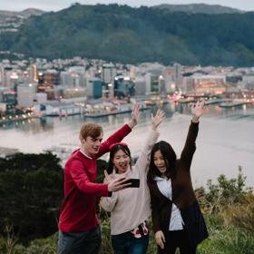Three people are taking a selfie at the Mount Victoria Lookout with a view of Wellington harbour and city centre behind them.