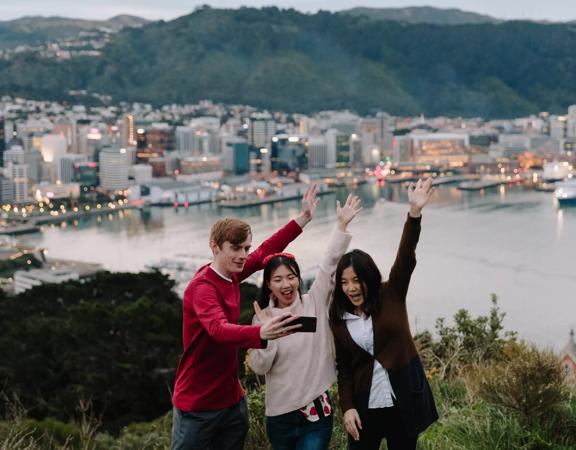 Three people are taking a selfie at the Mount Victoria Lookout with a view of Wellington harbour and city centre behind them.