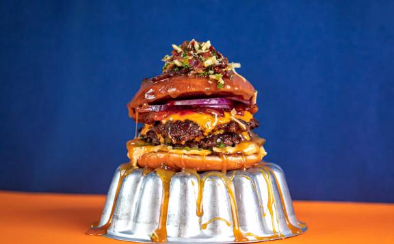 A donut bun filled with double-smashed patties and crispy bacon with cheese and salted caramel sauce drizzled down the sides sits on an upturned metal cake tin.