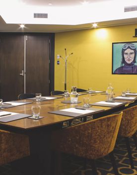 The interior of the Rita Angus Boardroom at the Bolton Hotel in Wellington with a large wood table, nine chairs and a dark yellow wall with a TV screen centrally mounted.