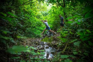 A person leaps across a stream while another follows, on the East harbour Regional park Kowhai Street track.