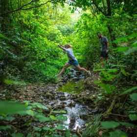 A person leaps across a stream while another follows, on the East harbour Regional park Kowhai Street track.