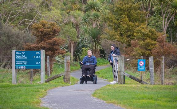 Two people on the ashphalt path on Te Ara o Tipapa trail - one is holding open the gate for a wheelchair user.