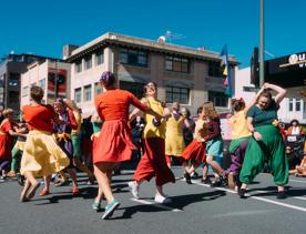 Dancers in colourful clothes pair up, hold hands and skip in circles.