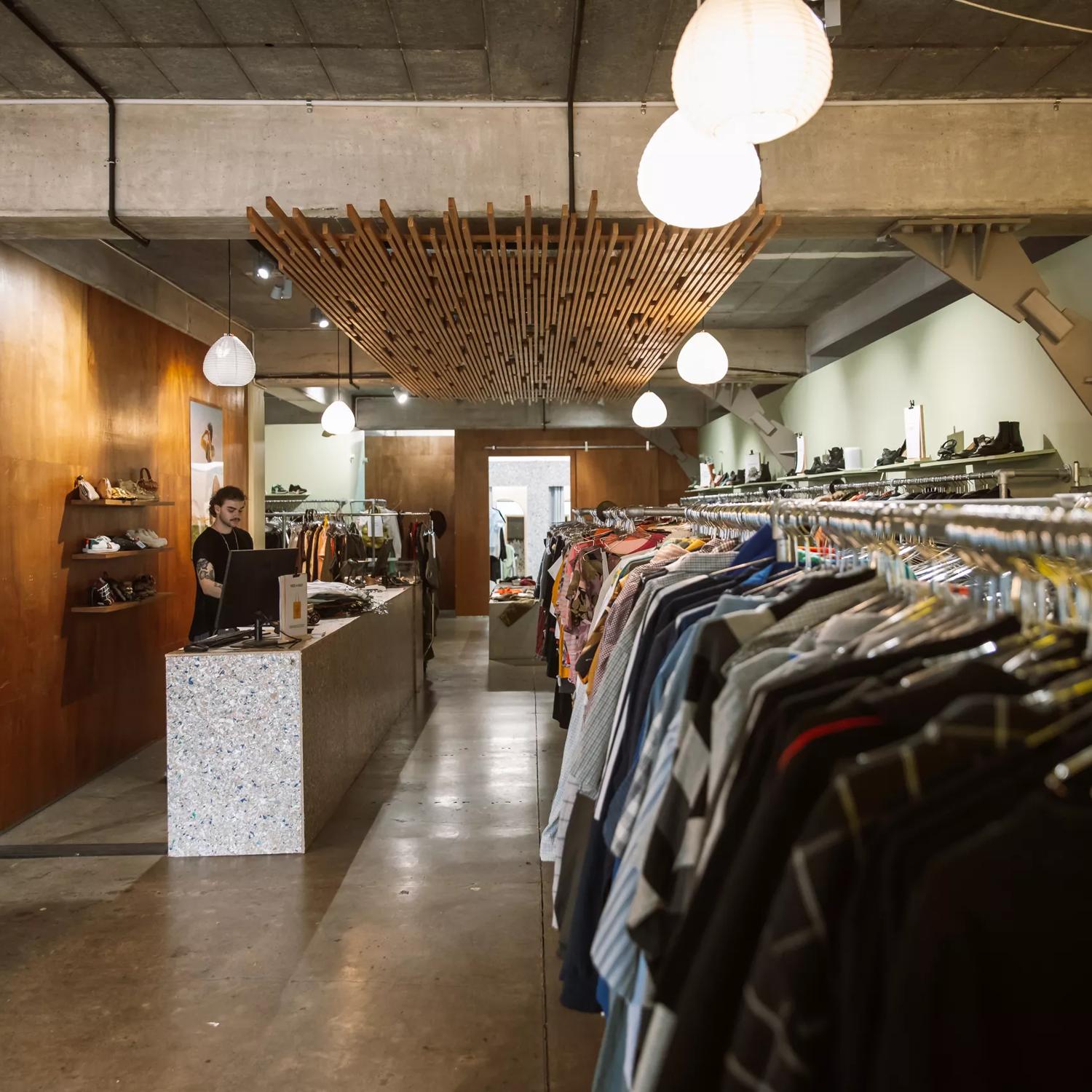 Inside Recycle Boutique, a secondhand clothing store on Ghuznee Street in Te Aro, Wellington. The space has concrete floors, ceilings, and wood-panelled walls. A worker wearing black is standing behind the counter.