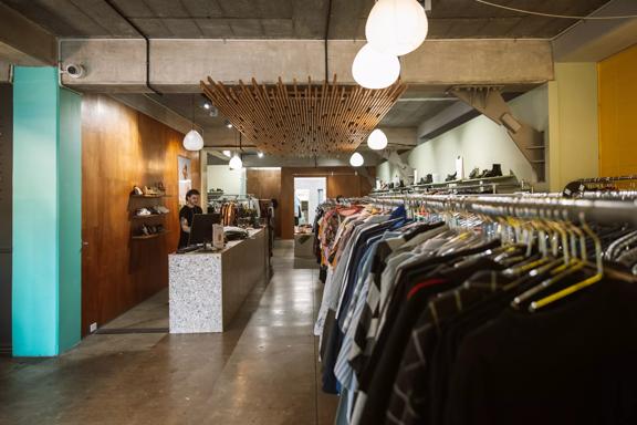 Inside Recycle Boutique, a secondhand clothing store on Ghuznee Street in Te Aro, Wellington. The space has concrete floors, ceilings, and wood-panelled walls. A worker wearing black is standing behind the counter.