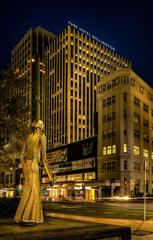 The Woman of Words sculpture stands in the foreground, with the Park Hotel stands in the distance. It is night and everything has a yellow glow.