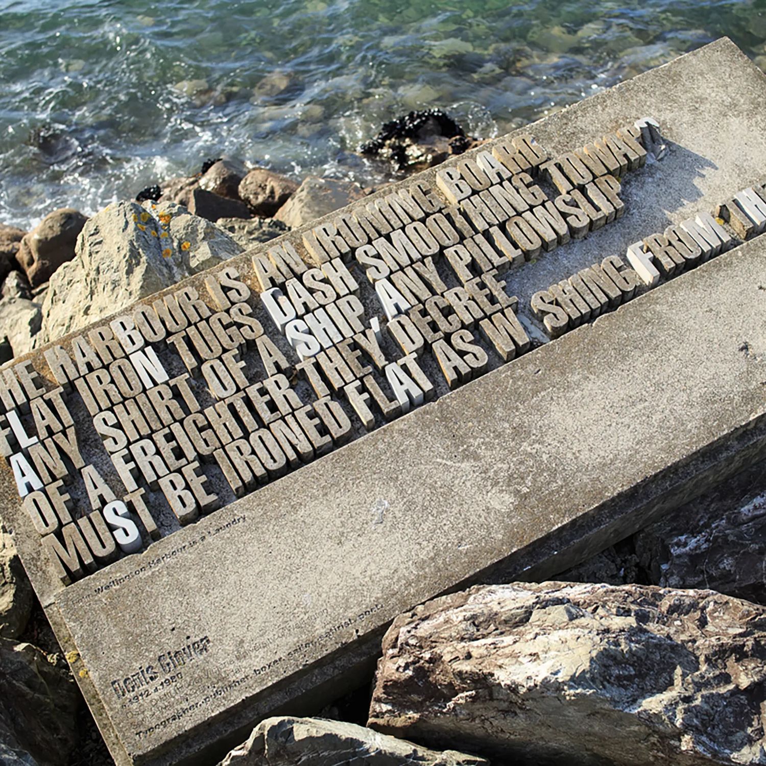 A typographical sculpture sits among rocks as the ocean laps at it. It says "The harbour is an ironing board flat iron tugs dash smoothing toward any shirt of a ship, any pillowslip of a freighter they decree must be ironed flat as washing from the sea." 
