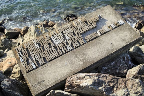 A typographical sculpture sits among rocks as the ocean laps at it. It says "The harbour is an ironing board flat iron tugs dash smoothing toward any shirt of a ship, any pillowslip of a freighter they decree must be ironed flat as washing from the sea." 
