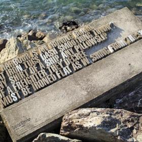 A typographical sculpture sits among rocks as the ocean laps at it. It says "The harbour is an ironing board flat iron tugs dash smoothing toward any shirt of a ship, any pillowslip of a freighter they decree must be ironed flat as washing from the sea." 
