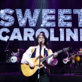 Singer playing guitar on stage with “SWEET CAROLINE” displayed behind.