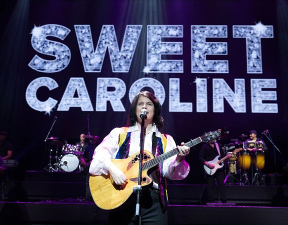 Singer playing guitar on stage with “SWEET CAROLINE” displayed behind.