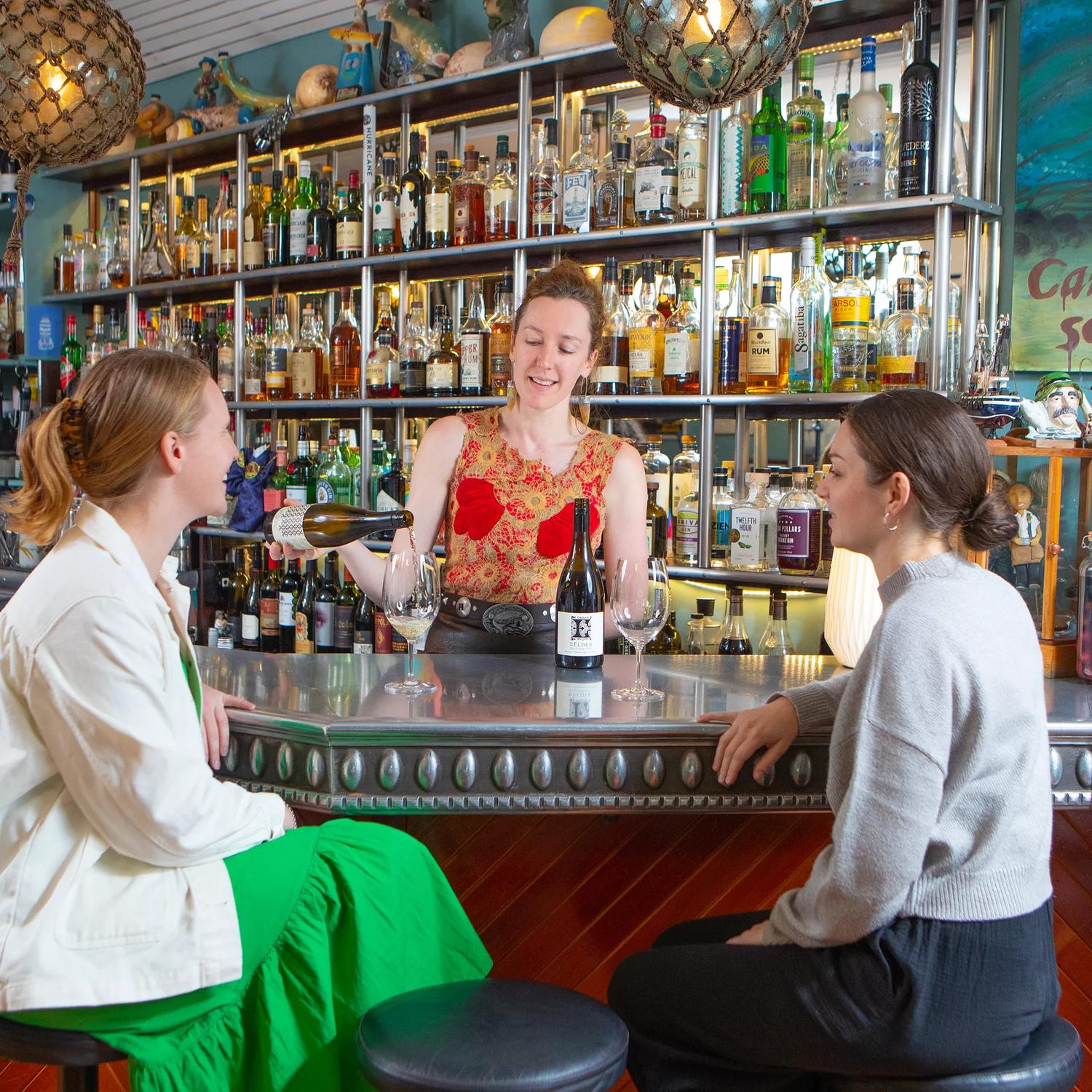 A bartender is pouring a glass of white wine for two people sitting at the bar at Ortega Fish Shack, a seafood restaurant in Wellington, New Zealand.