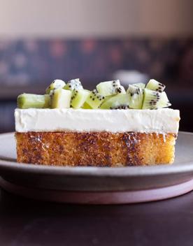 A slide of olive oil cake with cream cheese icing and kiwi fruit topping on a pale grey plate placed on a wooden table.