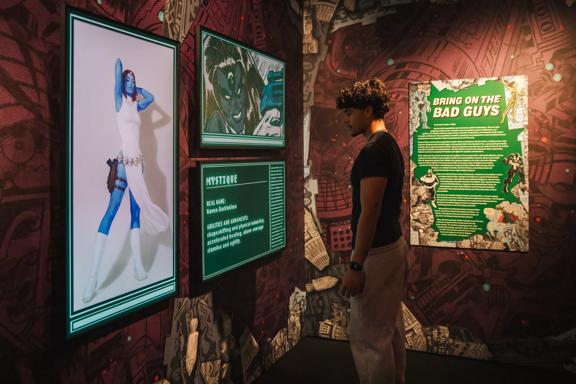 A person in a black T-shirt and grey pants  is looking at a panel about Mystique from X-Men at Marvel Earth's Mightiest Exhibition.