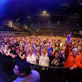 Looking over a large crowd of people inside TSB arena at a concert, the lights from above shine yellow red, and blue below.