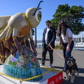 Decorative bee sculpture covered in colorful mosaic patterns displayed on a waterfront promenade, with two people and a dog standing beside it and boats in the background.