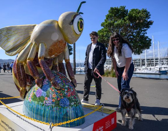 Decorative bee sculpture covered in colorful mosaic patterns displayed on a waterfront promenade, with two people and a dog standing beside it and boats in the background.