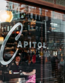 Looking into Capitol Restaurant from the outside, with the sign on the window. Two people enjoy a meal inside.