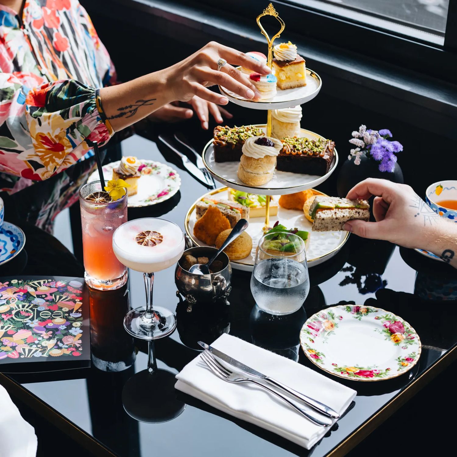 People enjoy high tea at QT Wellington. A three tiered plat, cocktails, and fancy teapots sit on the table.