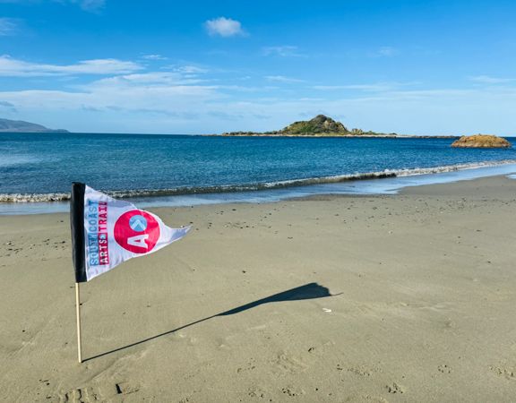 A flag planted in the sand on a quiet beach overlooks the ocean, with a small island visible offshore under a blue sky.