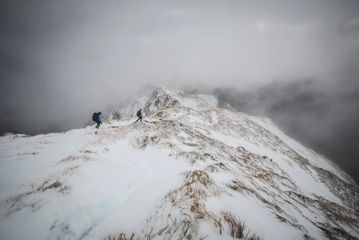Two hikers walking along a snowy mountain ridge, surrounded by low cloud.