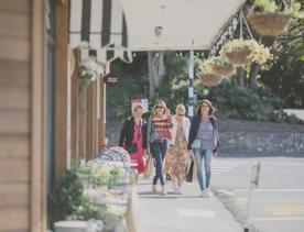 4 people walking along the footpath outside the Raumati shops. Above them are decorative lightbulbs and hanging planters.